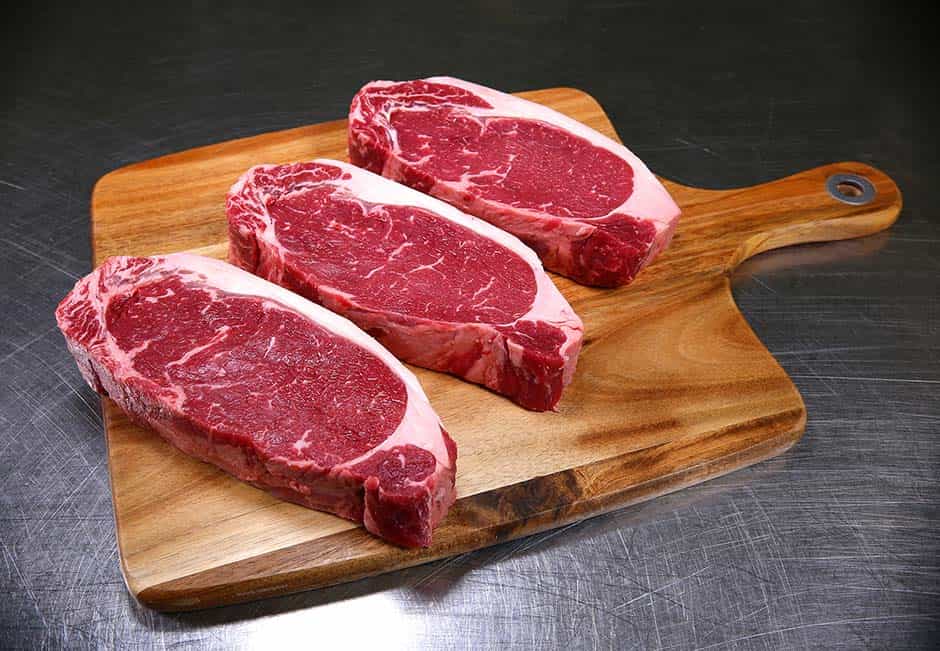 Tres bistecs de res crudos, incluidos cortes sirloin y ribeye, sobre una tabla